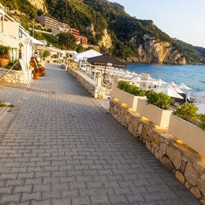 A coastal walkway with stone paving, outdoor seating, and umbrellas, leading to a beach area with a view of the sea and hills. - Achilleas Apartment  (Agios Gordios, Corfu)
