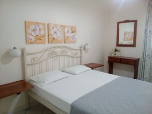 Une chambre à coucher avec un lit blanc, une table de chevet en bois, une lampe de chevet et un miroir au mur.