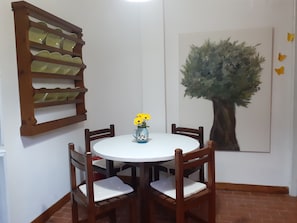 Un coin repas avec une table ronde, des chaises en bois, un vase avec des fleurs, une étagère fixée au mur et un tableau représentant un arbre.