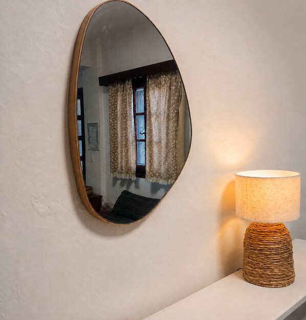 A mirror with a wooden frame on a wall, a lamp with a woven base, and a window with curtains.