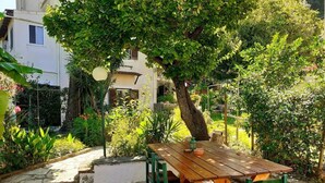 A wooden table with chairs in a garden area with stone flooring and various plants and trees. - Cottage House (Agios Gordios, Corfu)