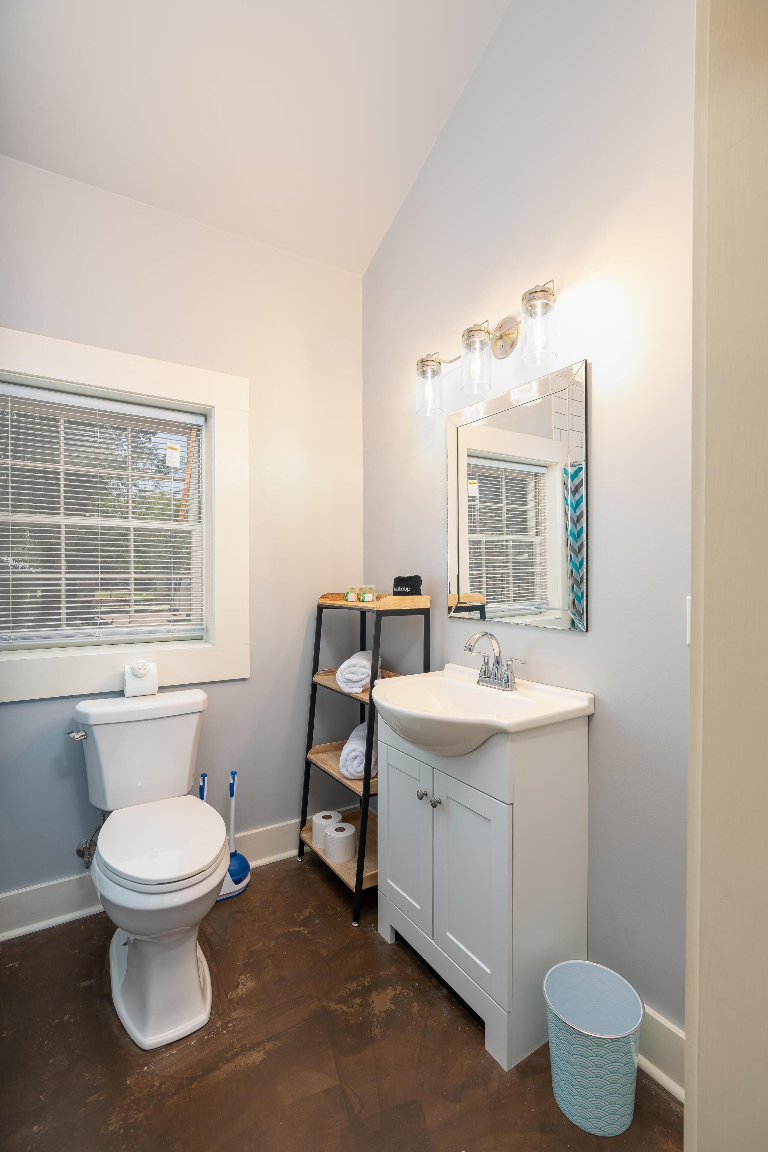 A bathroom with a toilet, sink, mirror, shelves, and a window.