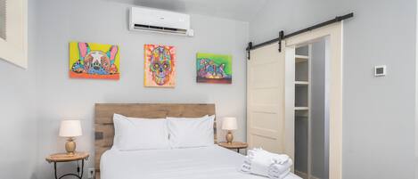 A hotel room with a wooden headboard, white bedding, two bedside tables with lamps, a closet with sliding doors, and three colorful paintings on the wall.