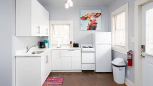 Cocina compacta con armarios blancos, estufa, pileta y basurero. Un tapete colorido está sobre el piso, y hay un cuadro de una vaca en la pared.