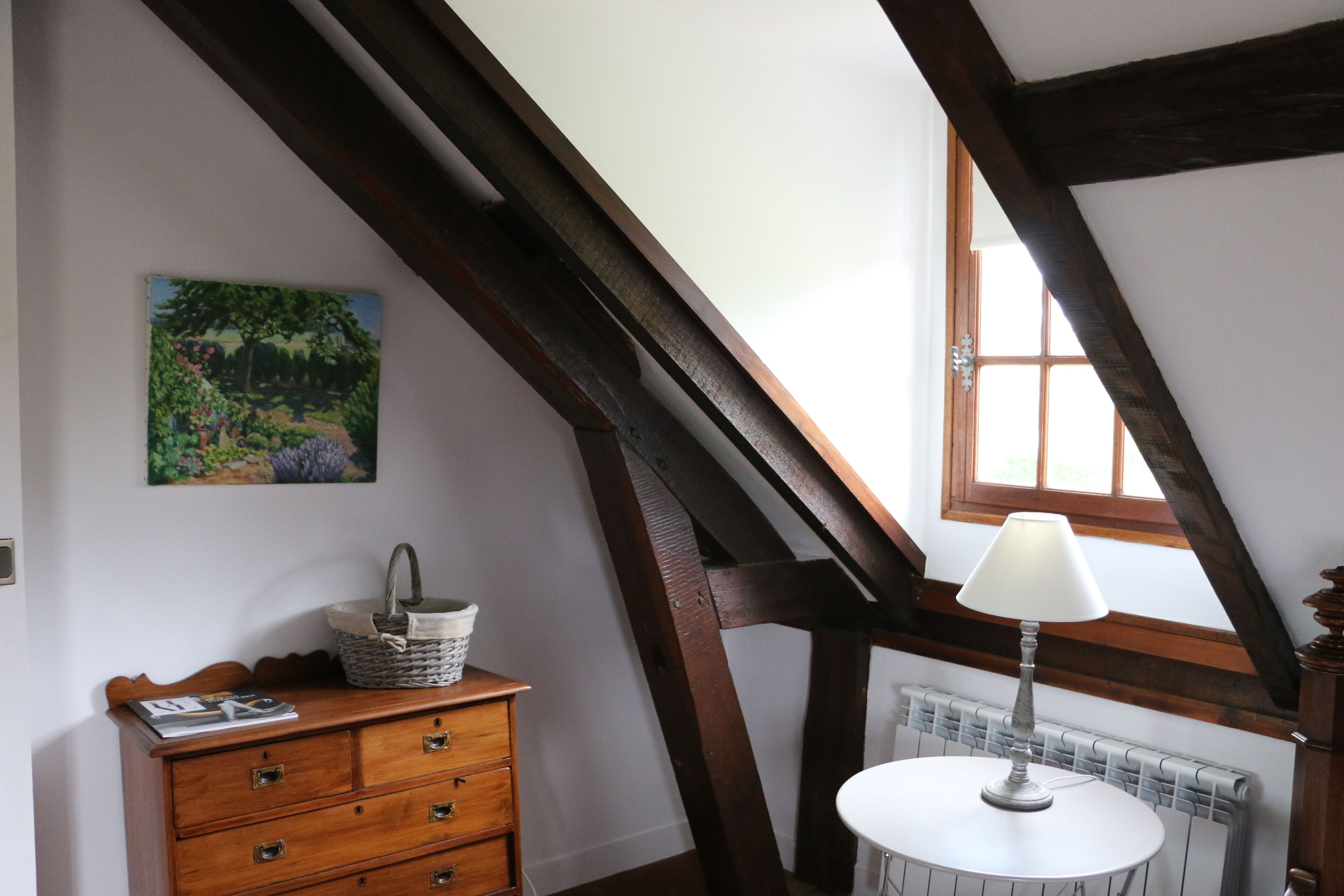 A room with a wooden dresser, a table with a lamp, a radiator, and a window with wooden beams.