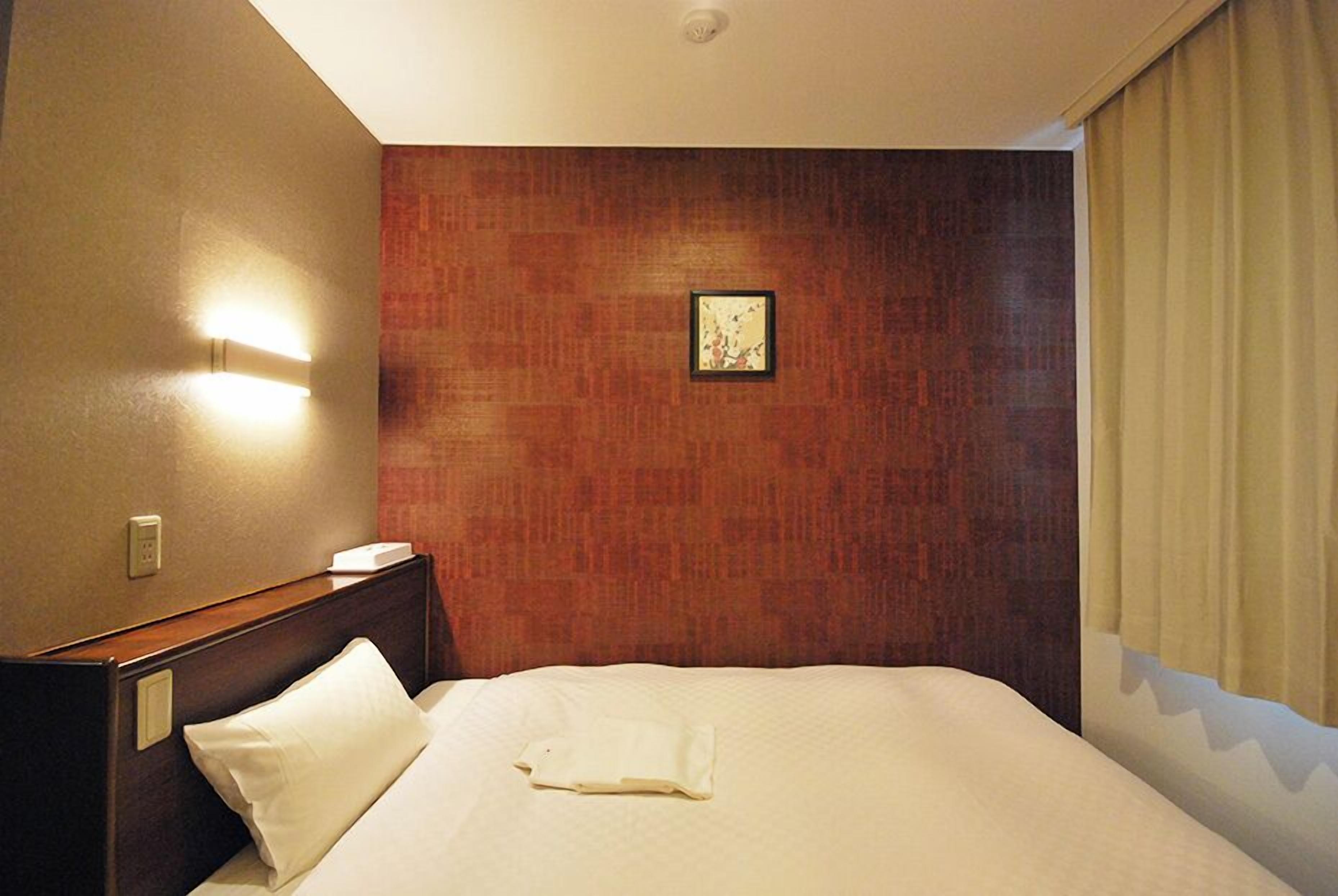 A hotel room with a neatly made bed, a headboard with a light switch, a framed picture on the wall, and a window with curtains.