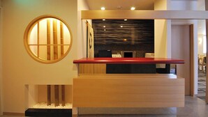 Un espace d’accueil avec un comptoir de réception en bois, une fenêtre ronde dotée de barreaux verticaux et un luminaire fixé au mur.
