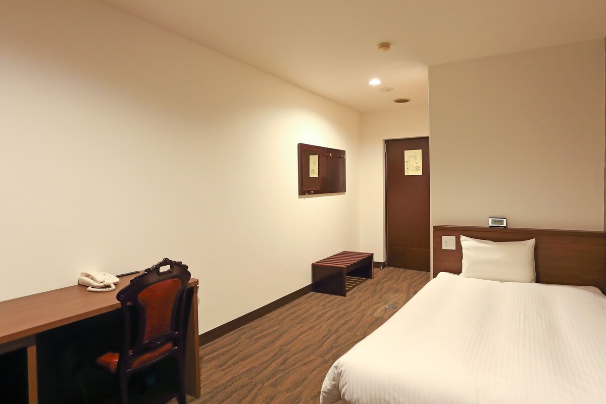 Een hotelkamer met een bed, een bureau met telefoon, een stoel, een bijzettafeltje en een wandklok.