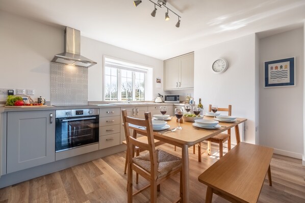 A modern kitchen with a built-in oven, a stainless steel range hood, and a dining area with a wooden table and chairs. - Twitchers Cottage, Titchwell, Norfolk (Titchwell)