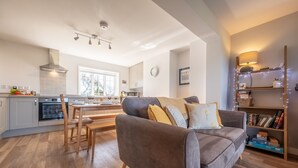 A modern living room with a grey sofa, wooden dining table, and a kitchen in the background. - Twitchers Cottage, Titchwell, Norfolk (Titchwell)