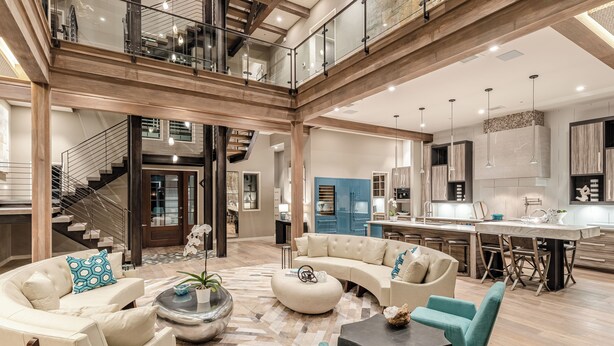 A spacious living area with a curved sofa, ottoman, and teal chairs. A staircase leads to an upper level with a glass railing. - Vermillion Sunsets | Lakefront Architectural Gem-Pool/Hot Tub/New 6 Seat LSV (Santa Rosa Beach)