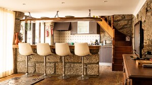 A kitchen with a stone wall, wooden bar stools, a wooden dining table, and a staircase leading to another level. - Casa da Eira - Casa Rural com Piscina Privada (Oliveira (São Pedro))