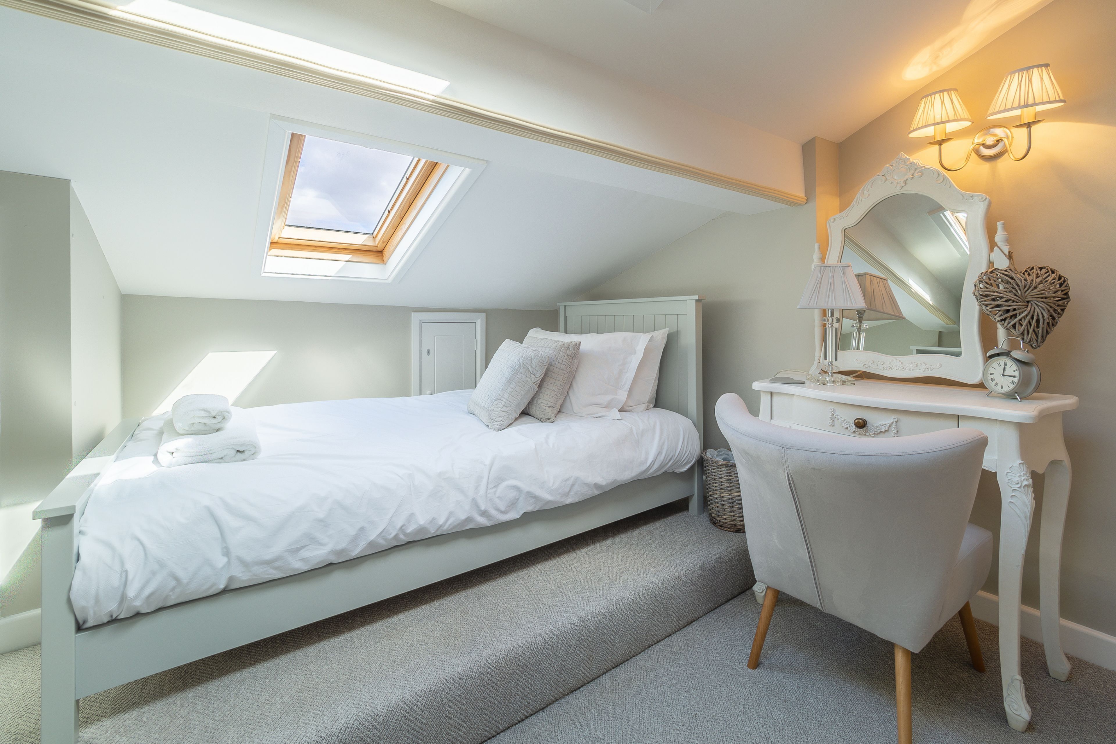 A bedroom with a skylight, a bed, a vanity with a mirror, and a chair.