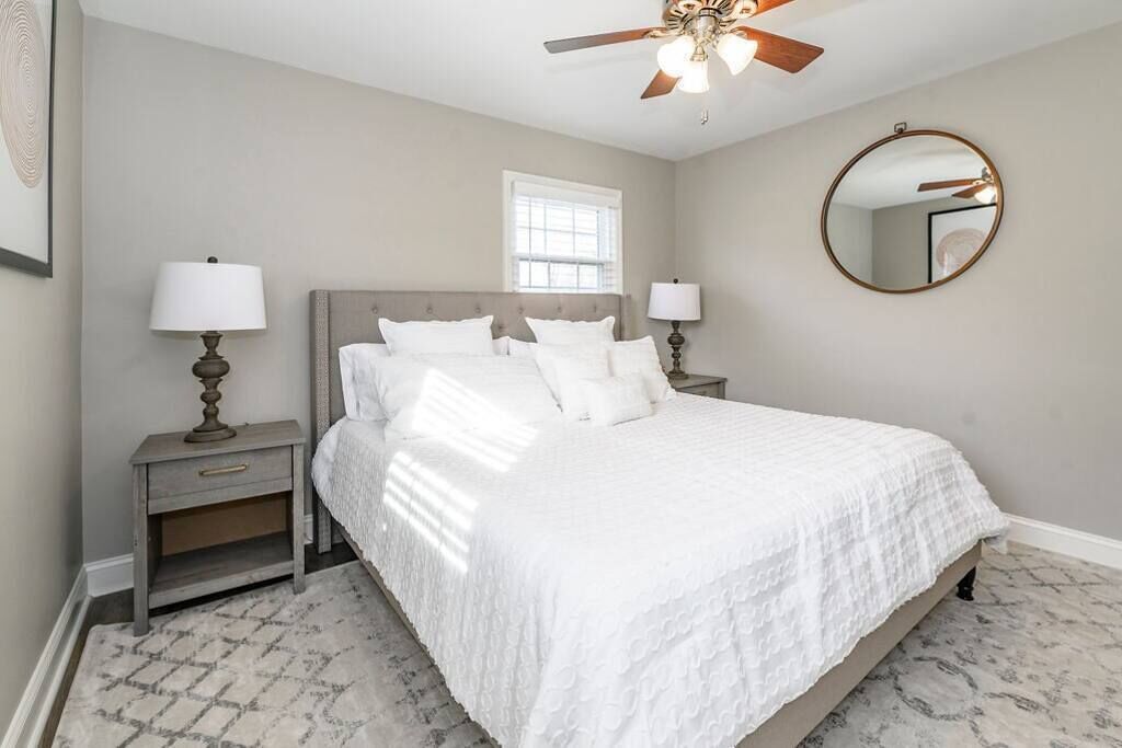 A bedroom with a bed, nightstands, a ceiling fan, a window, and a round mirror.