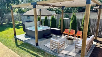 A hot tub under a wooden pergola with string lights, surrounded by outdoor furniture and a fire pit.