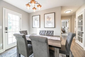 A dining room with a wooden table, four grey upholstered chairs, a patterned rug, and two framed leaf artworks on the wall.
