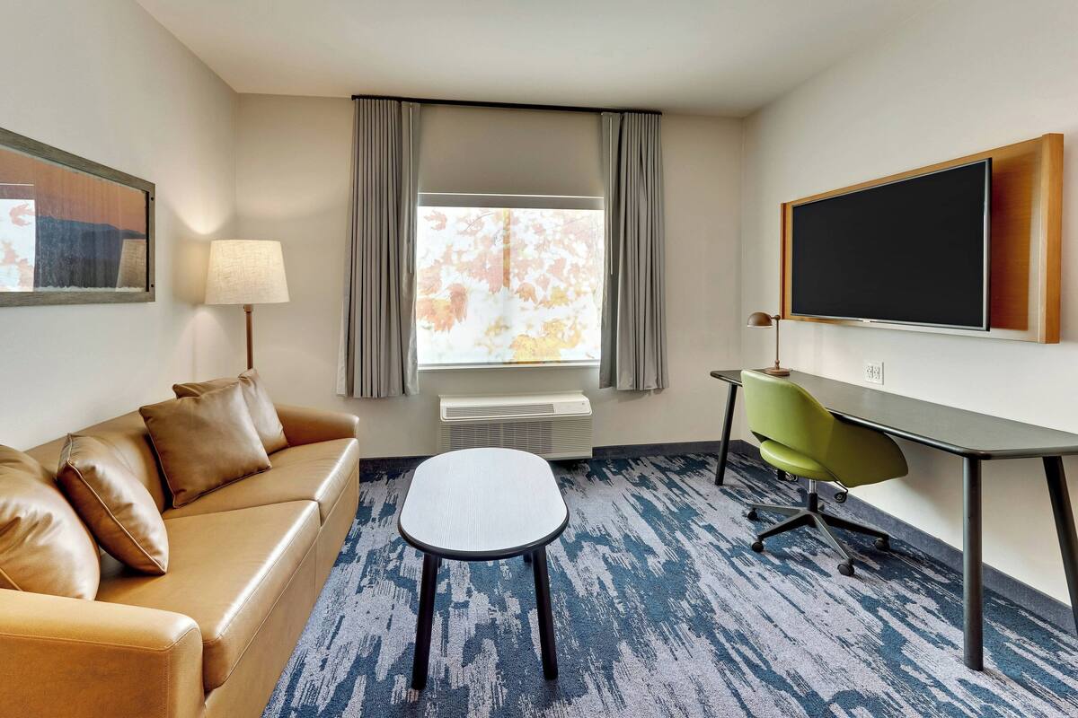 Habitación de hotel con sofá de cuero, una mesita pequeña, un escritorio con silla, televisor de pantalla plana, ventana con cortinas y un cuadro enmarcado en la pared.