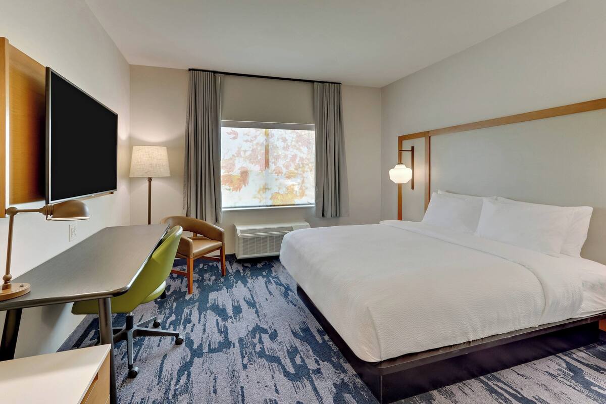 Habitación de hotel con cama, escritorio, silla, televisor y ventana con cortinas.