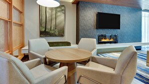 Un salon moderne avec une table ronde en bois, des fauteuils beiges, un téléviseur fixé au mur et une cheminée.
