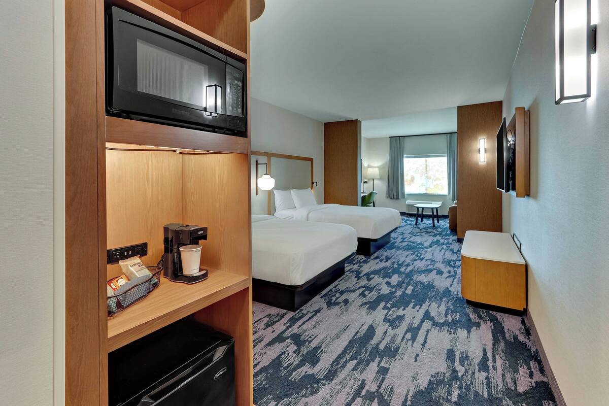 Habitación de hotel con microondas, cafetera y un refrigerador pequeño a la izquierda. Hay dos camas con ropa blanca y una mesita con una lámpara. La habitación tiene alfombra azul y una ventana con cortinas.