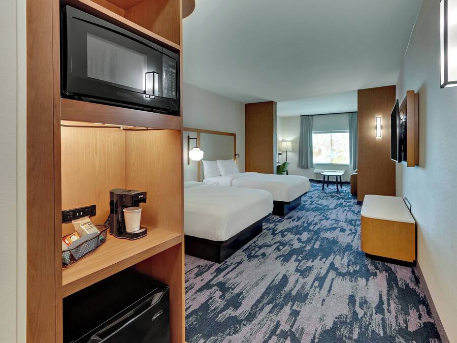 A hotel room with a microwave, coffee maker, and a small refrigerator on the left. There are two beds with white linens and a small table with a lamp. The room has a blue carpet and a window with curtains.