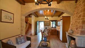 A spacious living area with a sofa, coffee table, and fireplace. A kitchen with wooden cabinets and a stone archway is visible in the background. - Christina II (,)