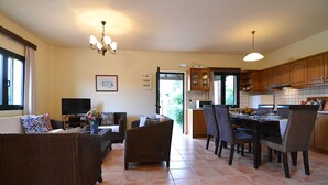 A spacious living area with a dining table and chairs, a television, and a kitchen in the background. - Christina III (Πενταμόδι, Κρήτη)