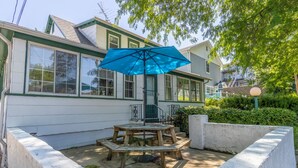 Une terrasse avec une table en bois et un parasol bleu, un bac à fleurs en béton, et une maison aux boiseries vertes et à la façade blanche.