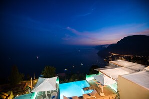 A rooftop pool area with lounge chairs and umbrellas, overlooking a coastal cityscape at night.