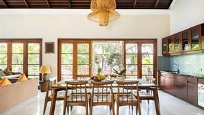 A spacious dining area with a wooden table and chairs, large windows with a view of greenery, and a modern kitchen with wooden cabinets.