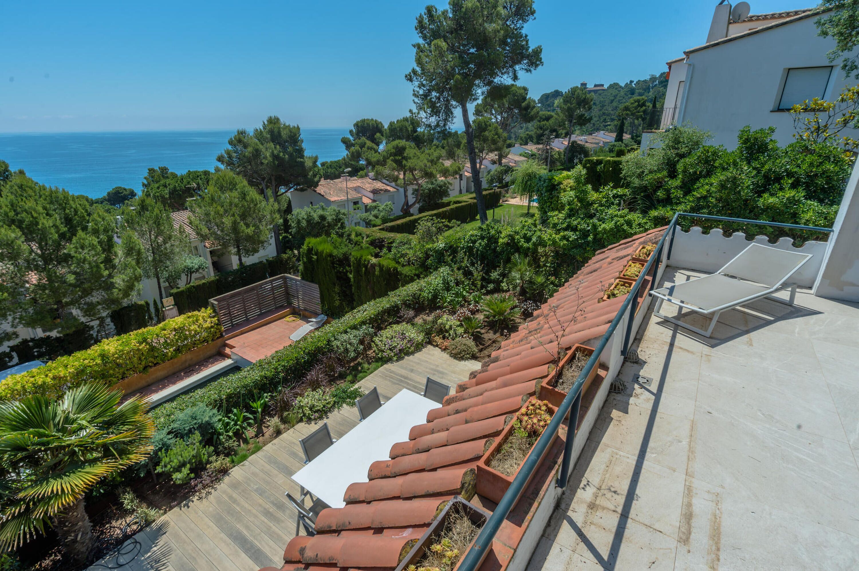 Terraço no telhado com mesa e cadeiras brancas, vasos de terracota e vista para o mar e vegetação circundante.