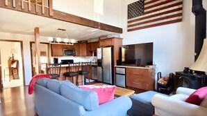 A spacious living area with a blue sofa, a red ottoman, and a kitchen in the background.