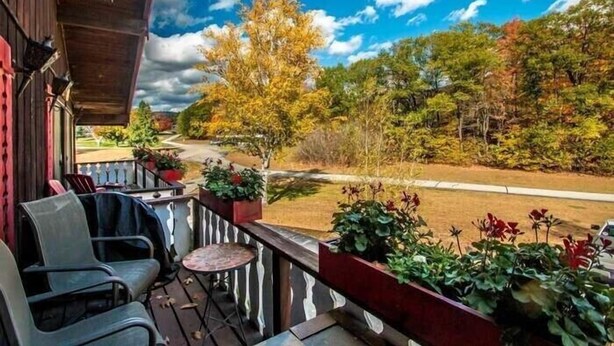 A wooden deck with outdoor seating and potted plants, overlooking a road and autumn foliage.