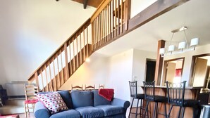 A spacious living area with a wooden staircase, a blue sofa set, a wooden coffee table, and a dining area with chairs.