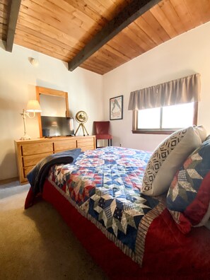 Een slaapkamer met een houten plafond, een bed met een quilt met patroon, een dressoir met een lamp, een stoel en een raam met gordijnen.