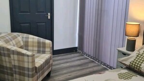 A hotel room with a plaid armchair, a bed with a floral bedspread, a dark door, and a window with vertical blinds. - Charming 1-bed Apartment in Peebles (Peebles)