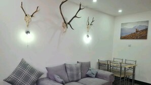 A living room with a grey sofa, plaid cushions, a red rug, and two mounted deer skulls with antlers. - Charming 1-bed Apartment in Peebles (Peebles)