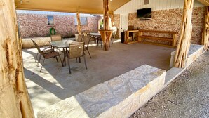 An outdoor patio area with a stone floor, wooden furniture, and a brick wall. - Concan River Ranch ~ 1,000' Private Frio River! Mtn. Views, Pet & Polaris OK. (Concan)