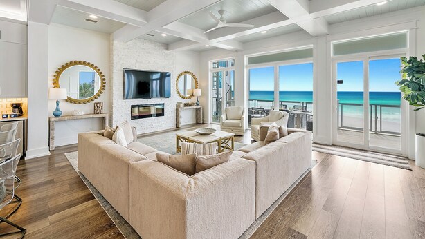 A spacious living room with a large beige sectional sofa, a flat-screen TV mounted on a white brick wall, and a balcony with a view of the ocean.