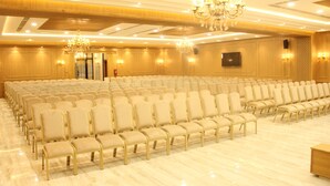 A large conference room with rows of beige chairs, a chandelier, and a flat-screen TV on the wall.