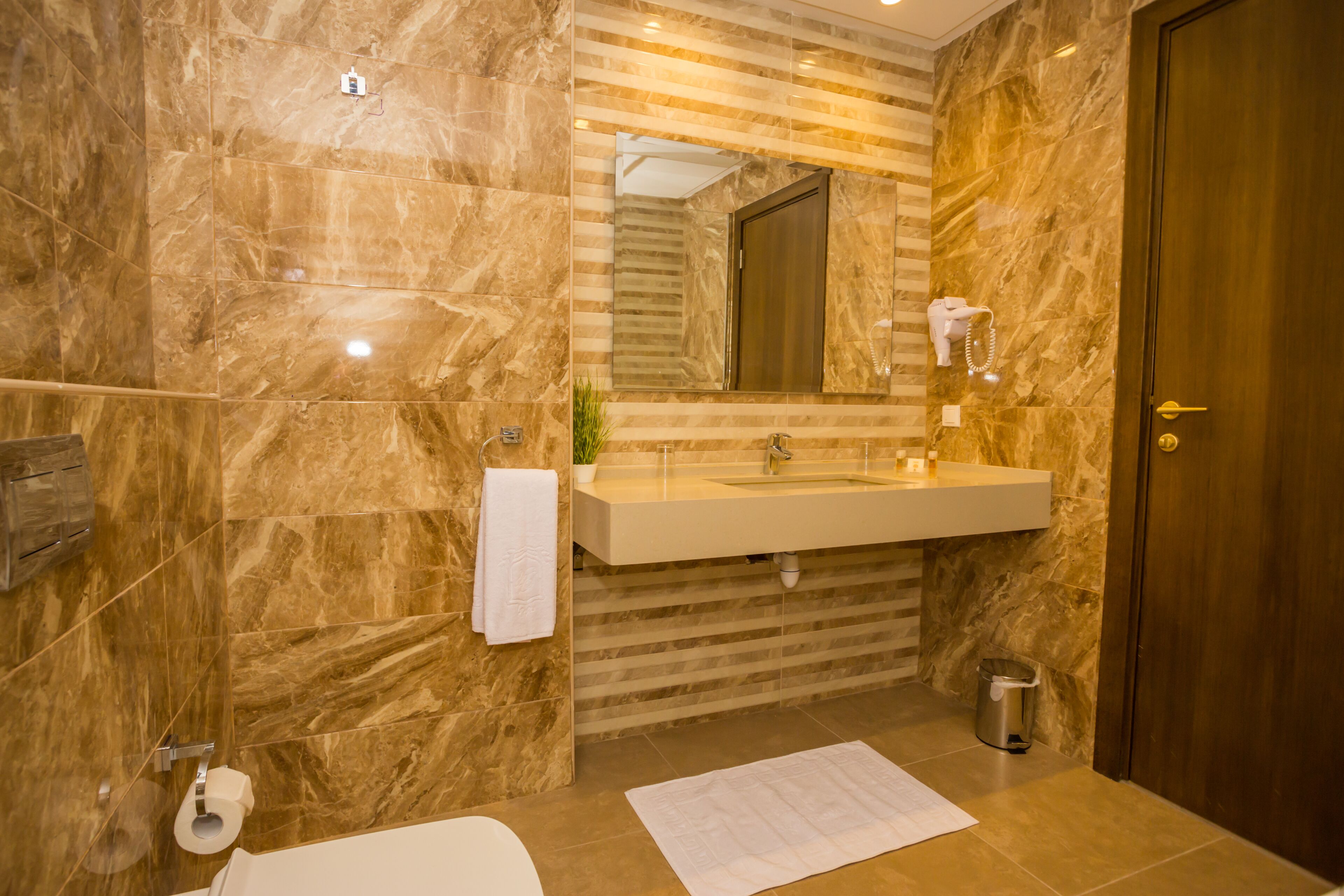 Un baño con pared de mármol, un lavamanos, un espejo, una toalla, un cubo de basura y una puerta.