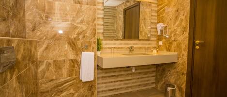 Un baño con pared de mármol, un lavamanos, un espejo, una toalla, un cubo de basura y una puerta.