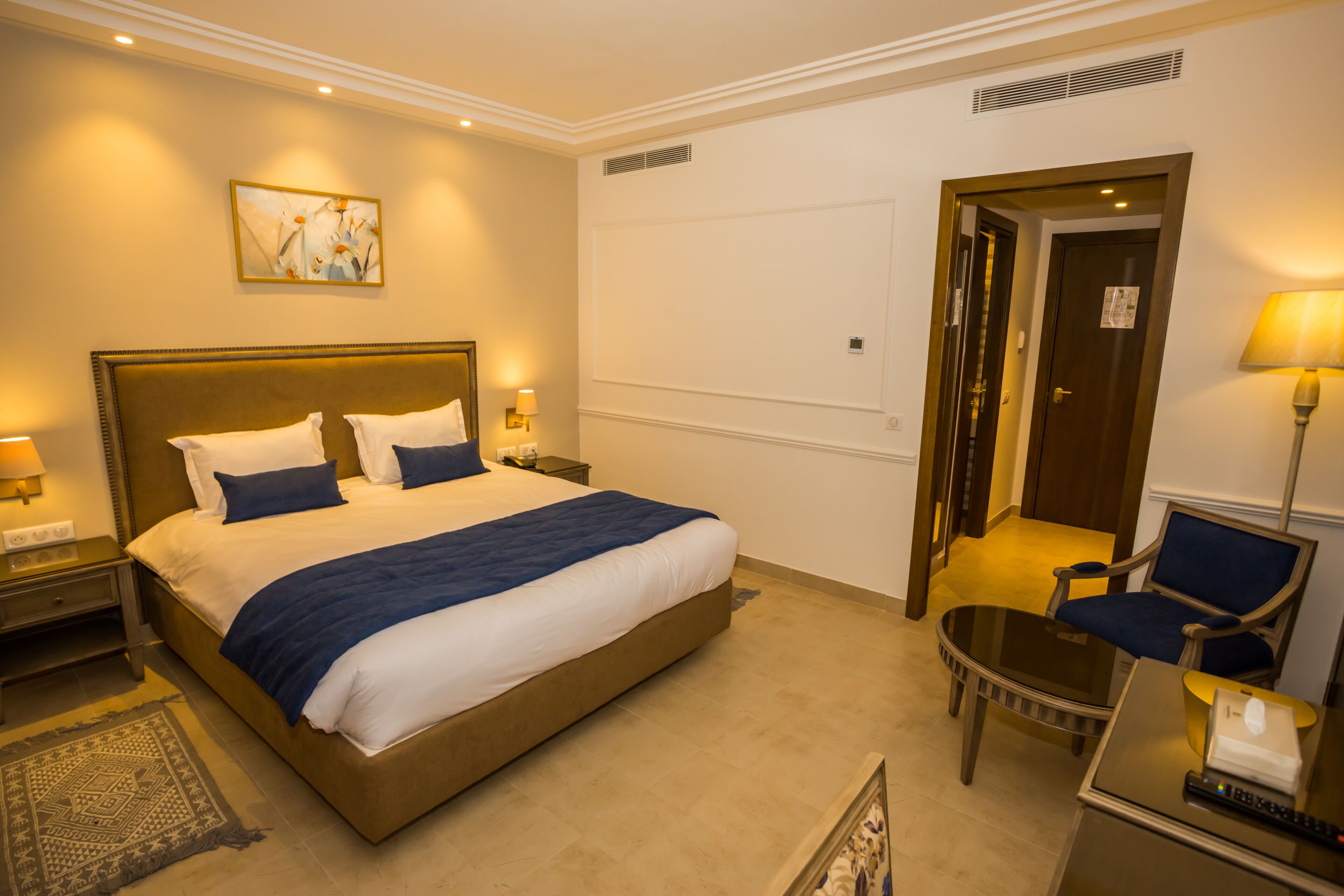 A hotel room with a large bed, bedside tables with lamps, a small table with a chair, and a door leading to another room.