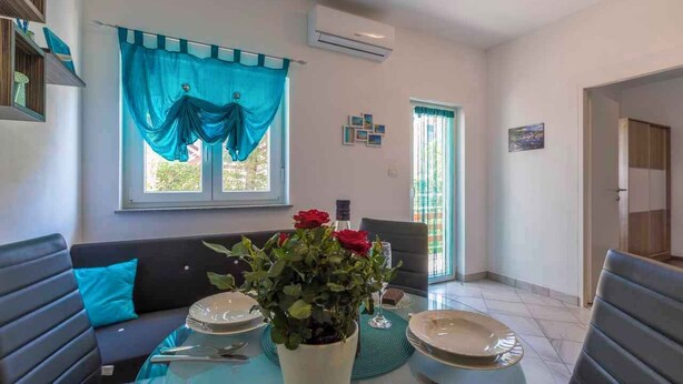 A dining area with a table set for two, a sofa with a blue cushion, and a window with blue curtains. - Holiday apartment with barbecue for shared use (Crikvenica)