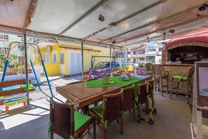 Un patio al aire libre con mesa y sillas de madera, una parrilla y una zona de juegos para niños con un tobogán.