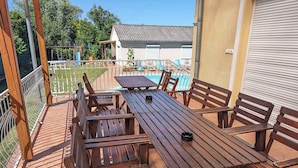 A wooden deck with a pool table and benches, surrounded by a metal fence and a view of a house in the background.