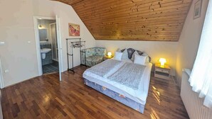A bedroom with a wooden ceiling, a bed, a nightstand, a wall-mounted drying rack, and a bathroom with a sink and toilet visible through an open door.