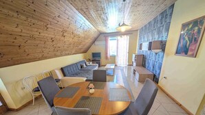 A cozy living room with a wooden ceiling, a sofa set, a dining table with chairs, and a TV.