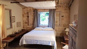 Een slaapkamer met een stenen muur, een bed met witte lakens, een houten bankje, een dressoir en een raam met gordijnen.