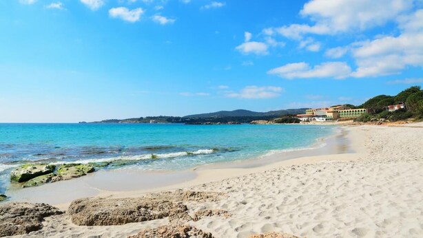A beach with clear blue water, sandy shore, and buildings in the background.